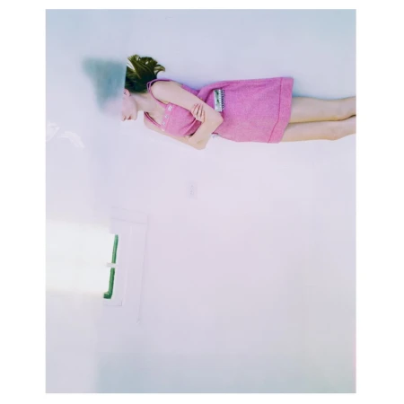 A woman lies on her side wearing a pink dress in a bright white room with a window. The image is high-key and in portrait orientation.