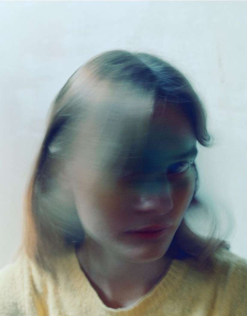 Portrait of a young woman with blurred motion effect, wearing a yellow sweater, set against a bright white background. Artful blur.