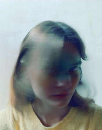 Portrait of a young woman with blurred motion effect, wearing a yellow sweater, set against a bright white background. Artful blur.