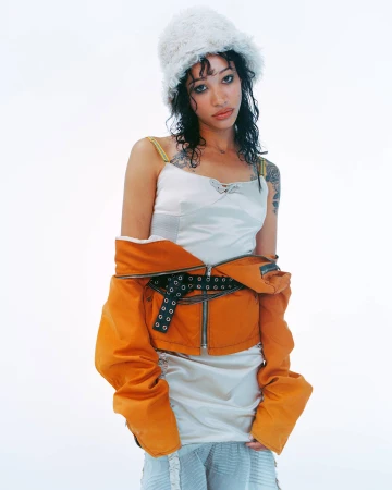 Woman in white slip dress and furry bucket hat wearing an orange jacket off her shoulders, against a white background.