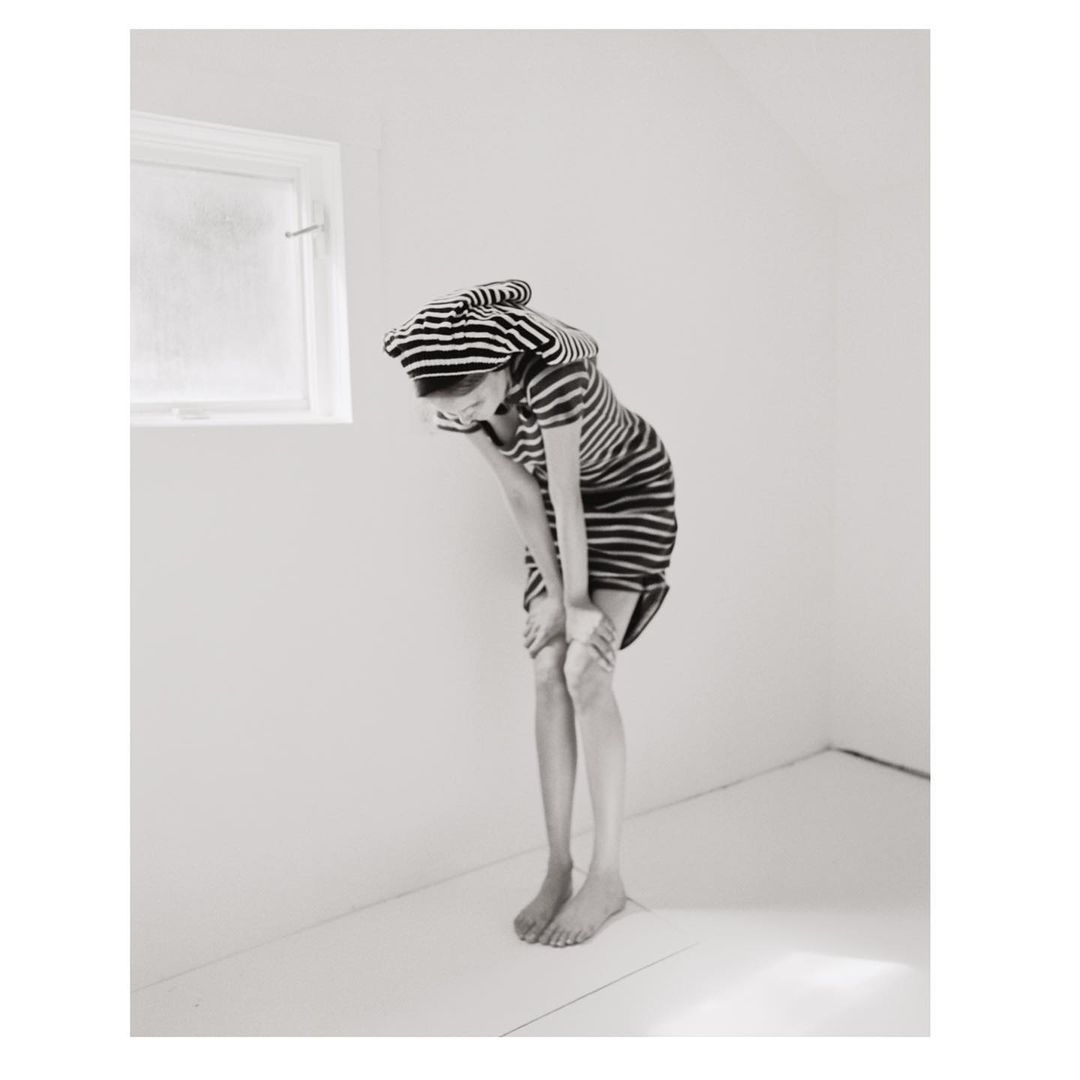 A child in a striped dress and cap stands barefoot in an all-white room, bending forward with hands on knees, near a small window.