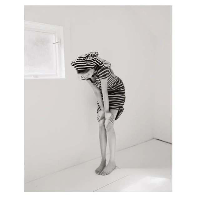A child in a striped dress and cap stands barefoot in an all-white room, bending forward with hands on knees, near a small window.