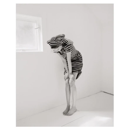 A child in a striped dress and cap stands barefoot in an all-white room, bending forward with hands on knees, near a small window.