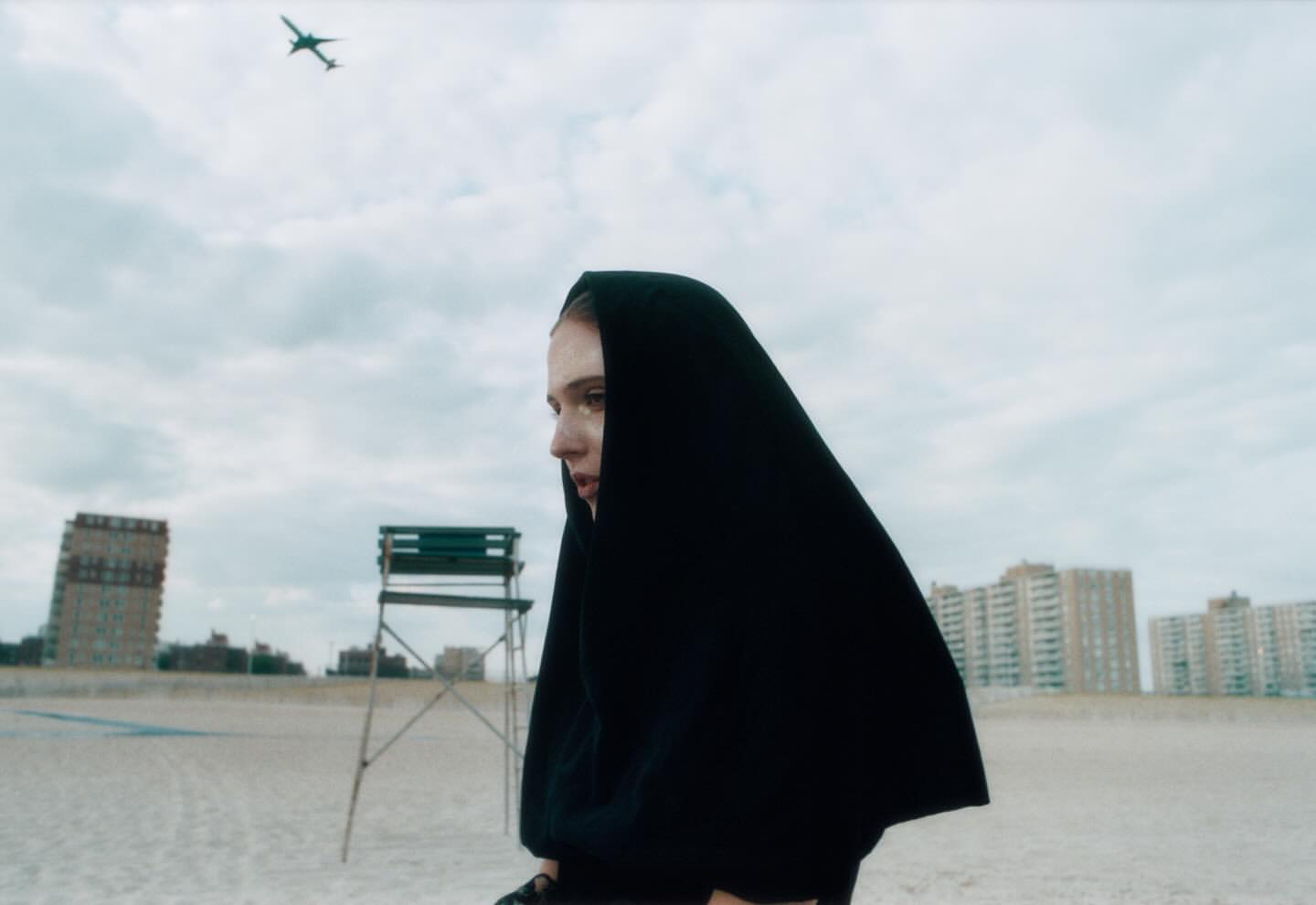 Woman in black hooded cloak on a beach with a distant cityscape and airplane overhead. Moody fashion editorial photography.