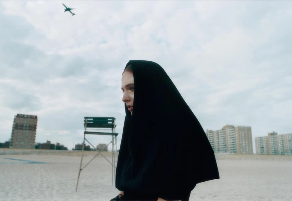 Woman in black hooded cloak on a beach with a distant cityscape and airplane overhead. Moody fashion editorial photography.