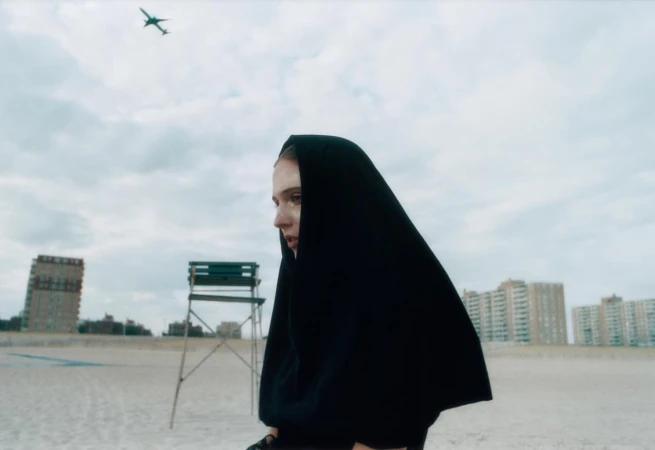 Woman in black hooded cloak on a beach with a distant cityscape and airplane overhead. Moody fashion editorial photography.