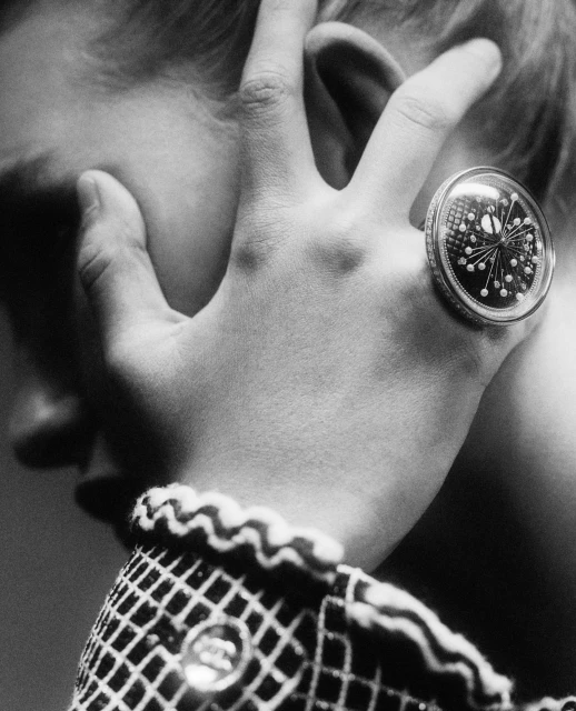 Close-up black and white photo of a person's hand with a large cocktail ring touching their ear, featuring a jacket with a grid pattern.