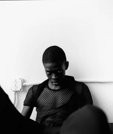 Monochrome portrait of a young adult, head and shoulders, wearing a mesh top, against a plain white wall with electrical outlet.