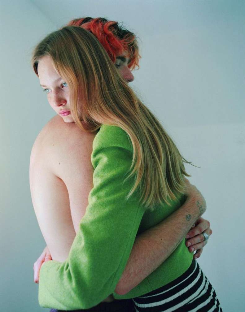 A blonde woman in a green jacket hugs a shirtless man with red-orange hair against a light blue wall, displaying an intimate moment.