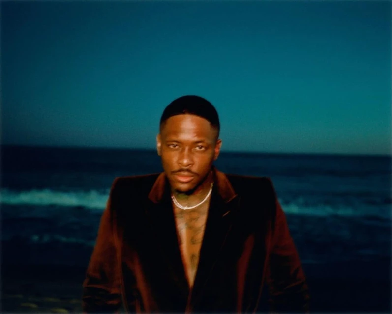 Portrait of an adult male wearing a dark brown suit and a white necklace standing in front of a dark blue ocean backdrop.