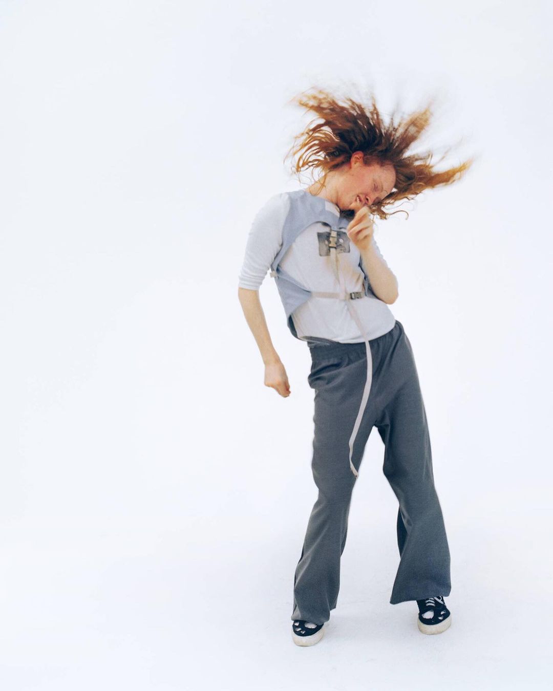 Young woman with red hair dynamically poses in gray pants, a white shirt, and a light gray vest against a stark white backdrop in a studio.