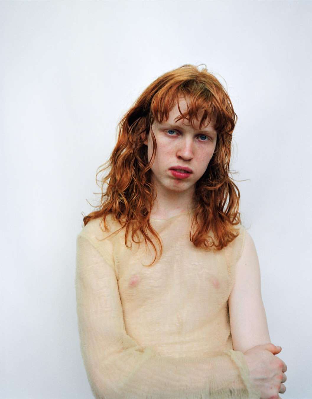 Portrait of a young adult with red hair and a sheer top standing against a white backdrop in a studio environment.