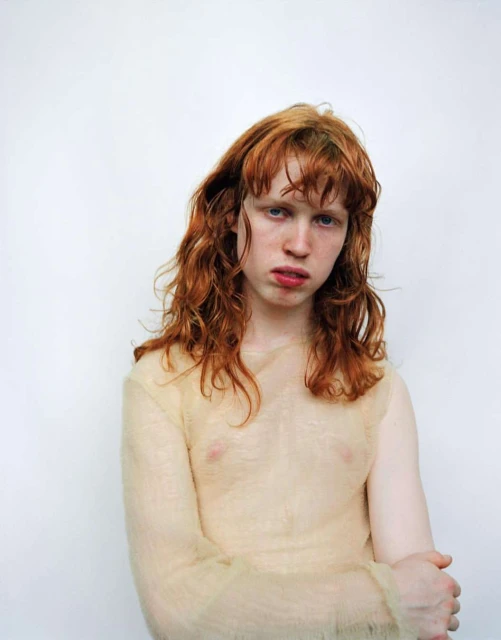 Portrait of a young adult with red hair and a sheer top standing against a white backdrop in a studio environment.