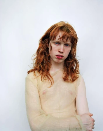 Portrait of a young adult with red hair and a sheer top standing against a white backdrop in a studio environment.