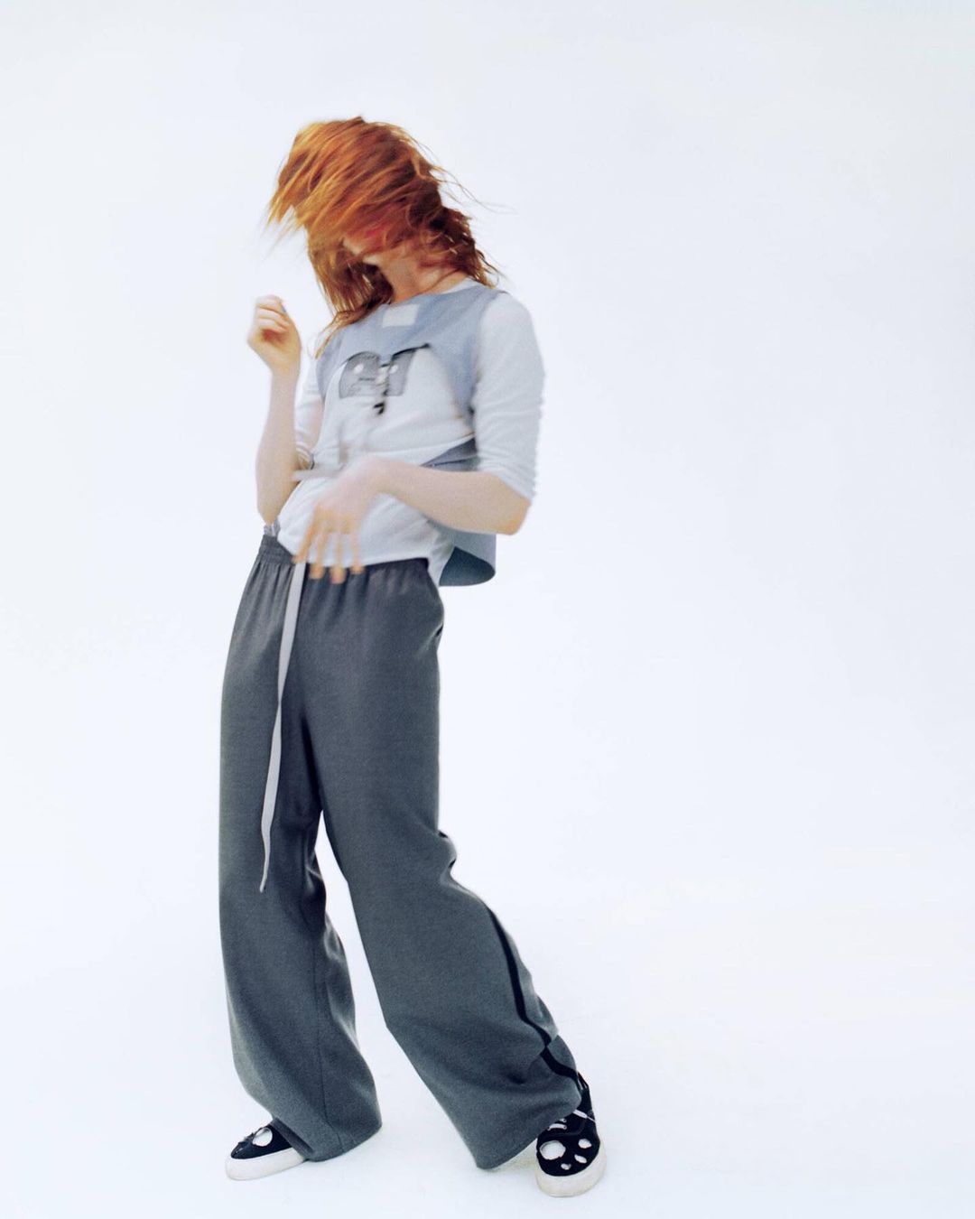 A person with red hair poses in a light gray top and slate-gray pants, with the figure slightly tilted back against a white background.