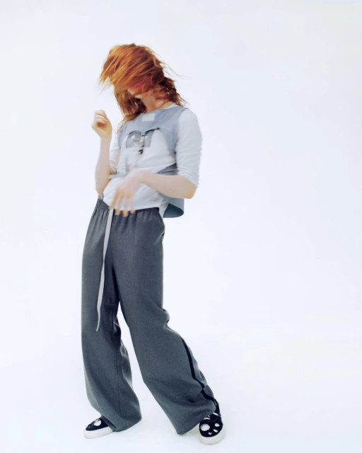 A person with red hair poses in a light gray top and slate-gray pants, with the figure slightly tilted back against a white background.