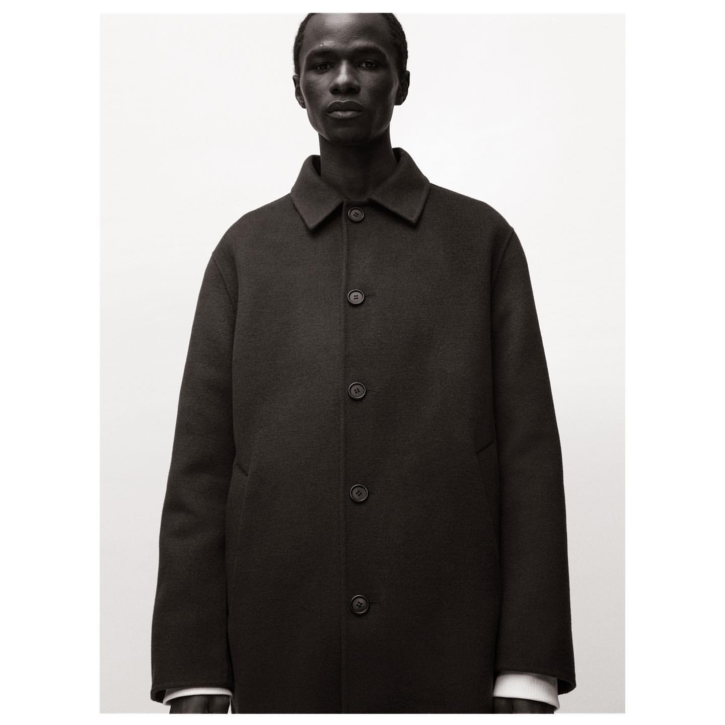 Headshot of a dark-skinned male model in a buttoned black wool overcoat against a neutral background in studio lighting.
