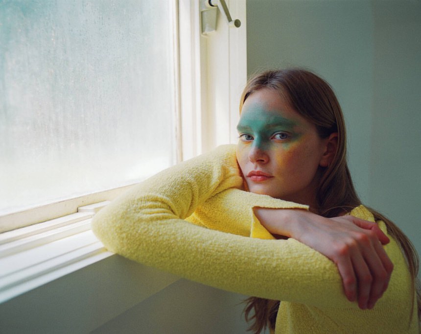 Young woman with creative face paint poses near a window in a serene indoor setting, highlighted by natural light and pastel colors.