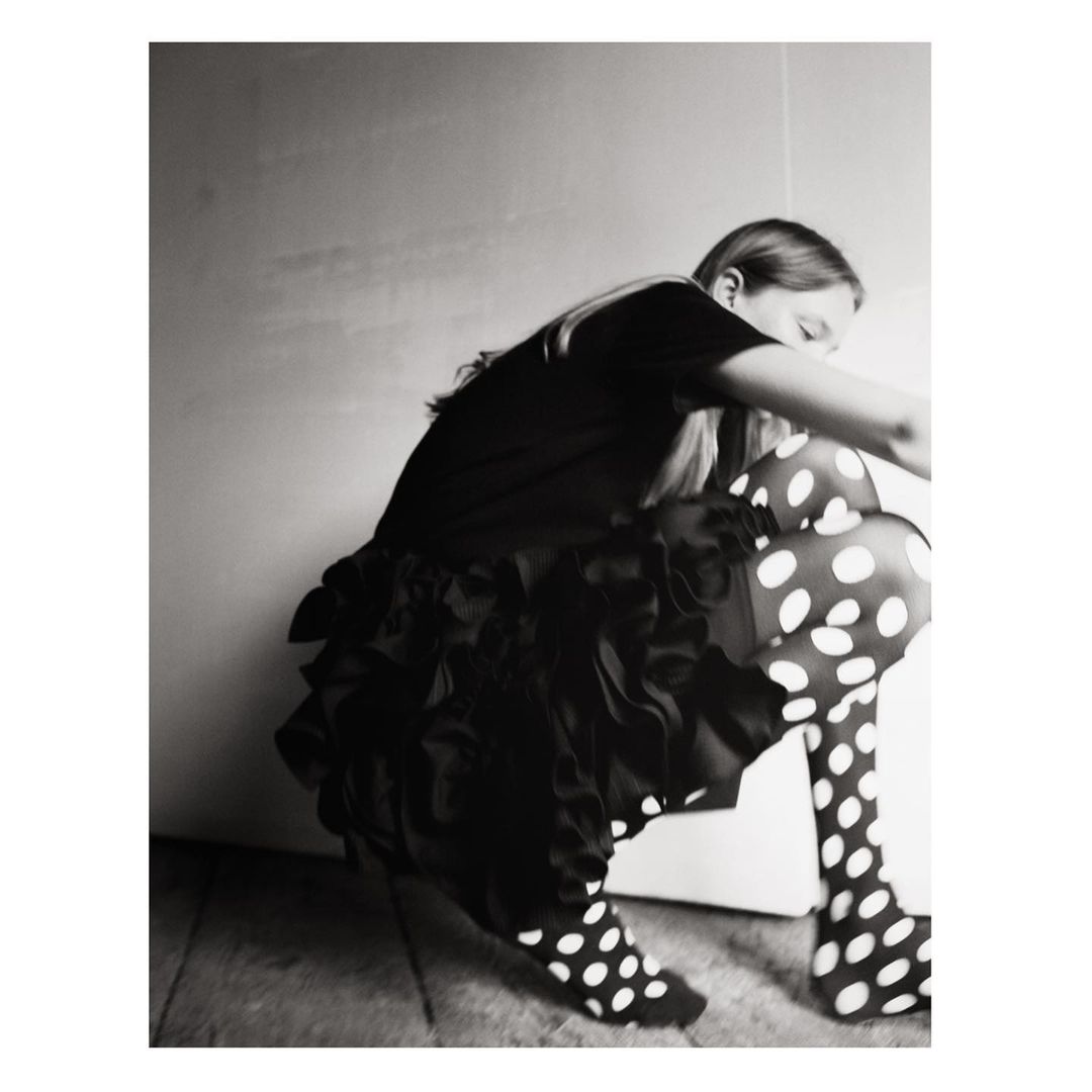 A girl with long blonde hair is crouching, wearing a black top, ruffled skirt, and polka dot tights, in a black and white studio shot.
