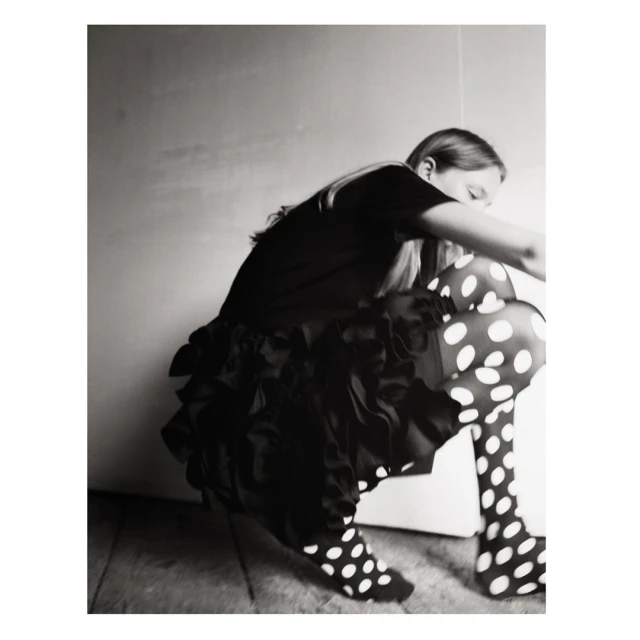 A girl with long blonde hair is crouching, wearing a black top, ruffled skirt, and polka dot tights, in a black and white studio shot.