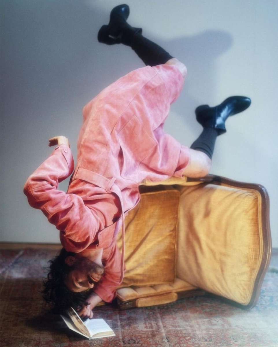 Person in pink coat and black boots playfully poses upside down on vintage luggage while reading a book indoors, artistic and whimsical.