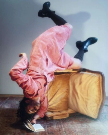 Person in pink coat and black boots playfully poses upside down on vintage luggage while reading a book indoors, artistic and whimsical.