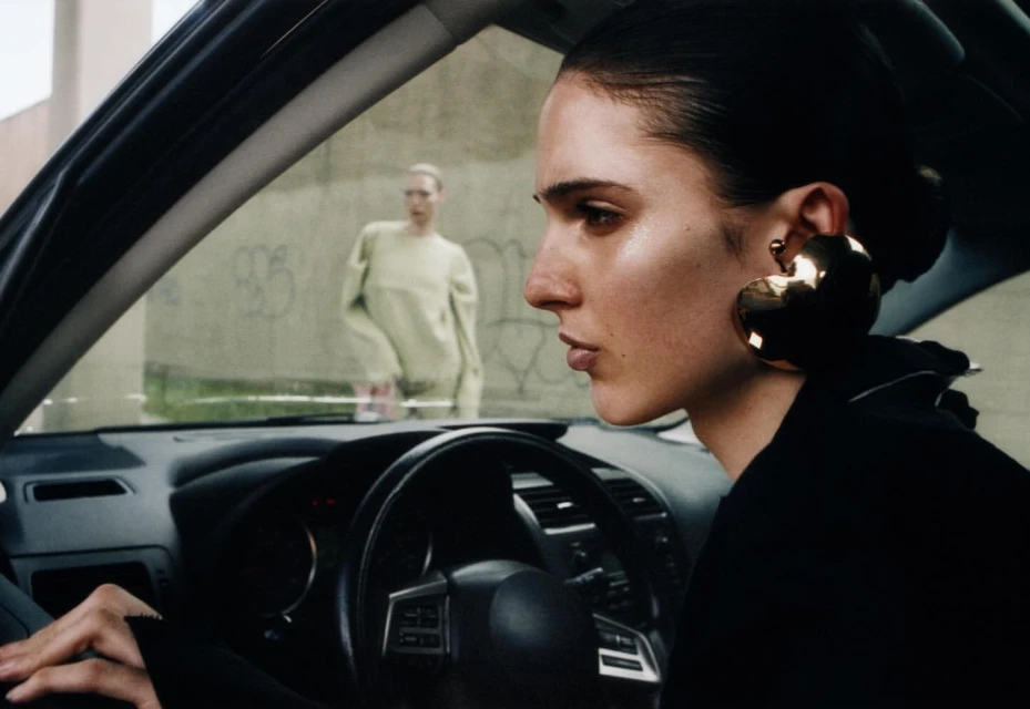 Two women pose next to a car; one is in the driver's seat wearing large gold earrings, and the other stands outside near a wall.