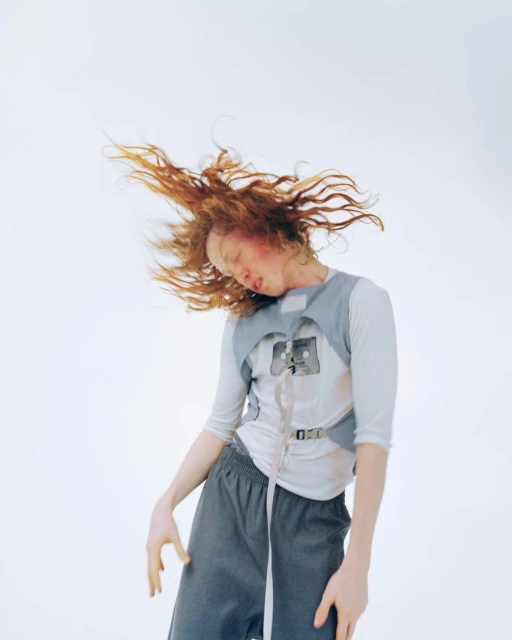 Red-haired person with eyes closed and head tilted, wearing a gray vest and loose pants against a white backdrop in a studio.