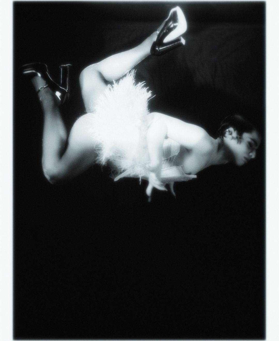 Monochrome portrait of reclining adult in white feathered clothing and high heels on a dark backdrop, low angle and close up.