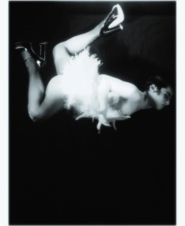Monochrome portrait of reclining adult in white feathered clothing and high heels on a dark backdrop, low angle and close up.