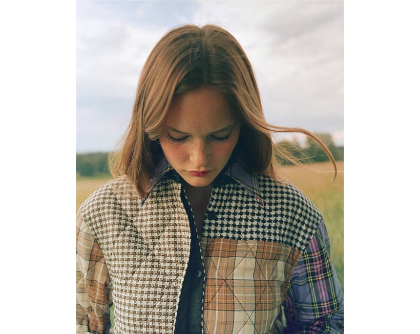 Young woman with long brown hair stands outdoors wearing a patchwork jacket in a field with a blurred background of trees and sky.