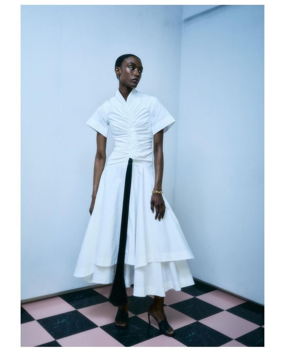 A single Black woman in a white dress with a black accent piece, posing in a corner with a checkered floor for an indoor fashion shot.