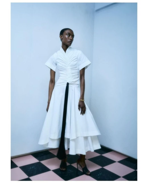 A single Black woman in a white dress with a black accent piece, posing in a corner with a checkered floor for an indoor fashion shot.