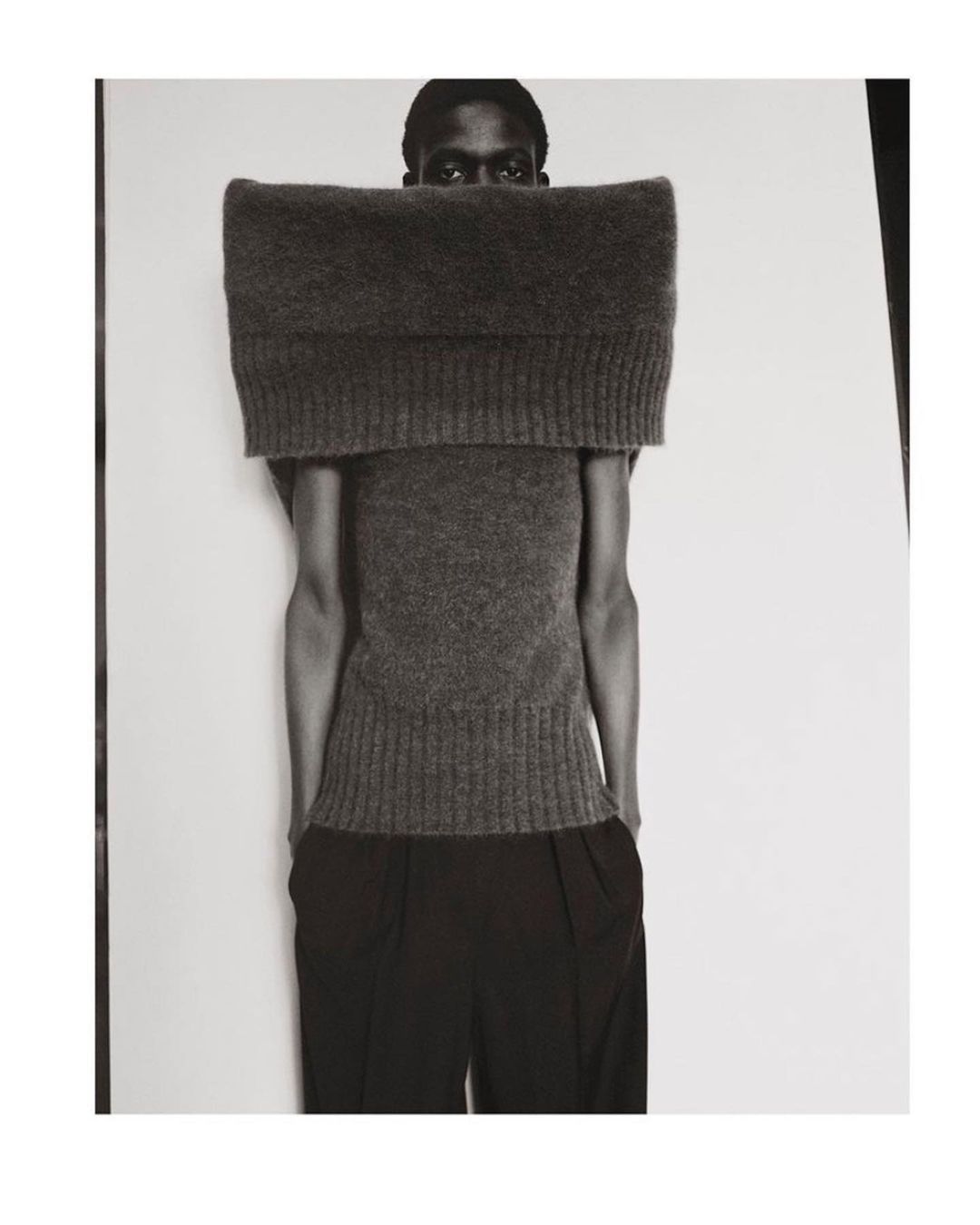 Monochrome studio shot of a dark-skinned adult model with eyes visible above an oversized grey knit top, and black trousers against a white background.