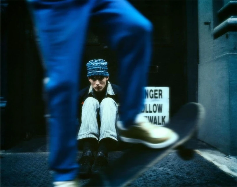 A teenager with a knitted hat sits near an entrance, while a skateboarder partially obstructs the view in an urban setting.