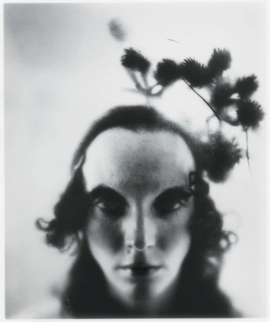 Monochromatic close-up of a person's face with dark hair and striking features, adorned with a delicate floral headpiece against a soft background.