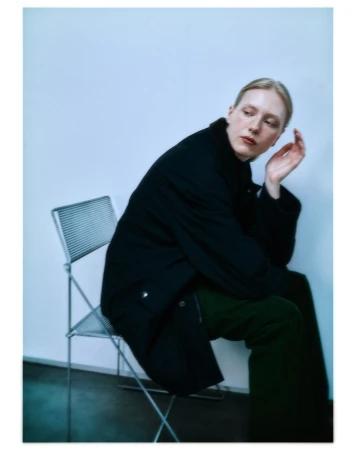 Blonde woman wearing a dark coat and green trousers seated on a wire chair against a white wall.