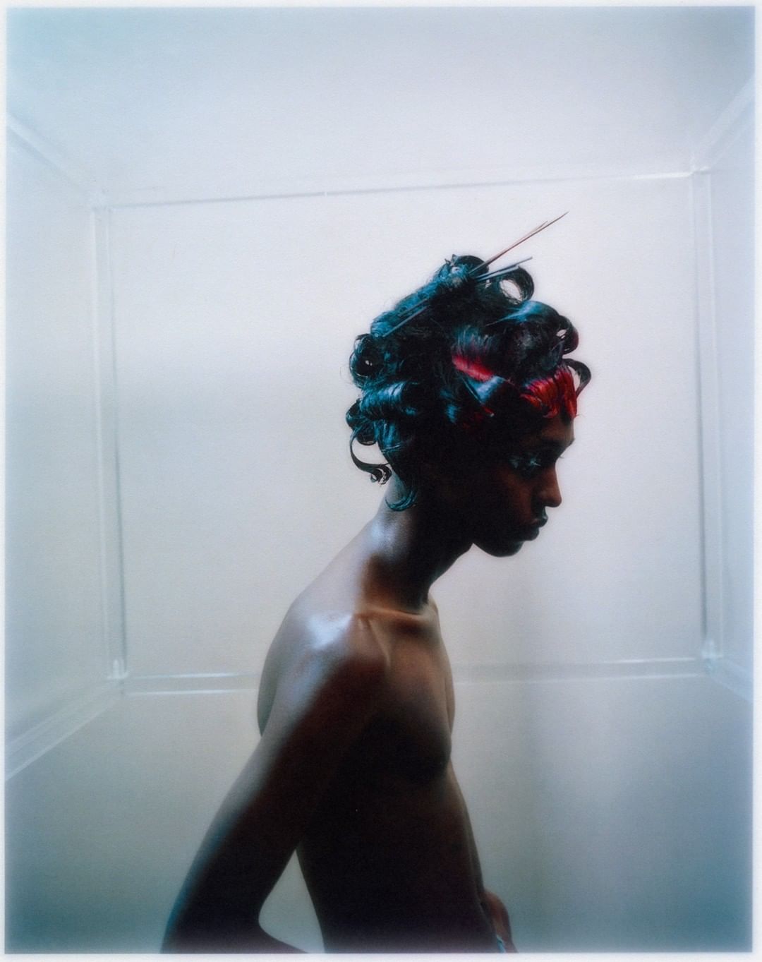 A person with dark skin and curly teal hair stands in a translucent white box, captured in a side-view studio shot with soft lighting.