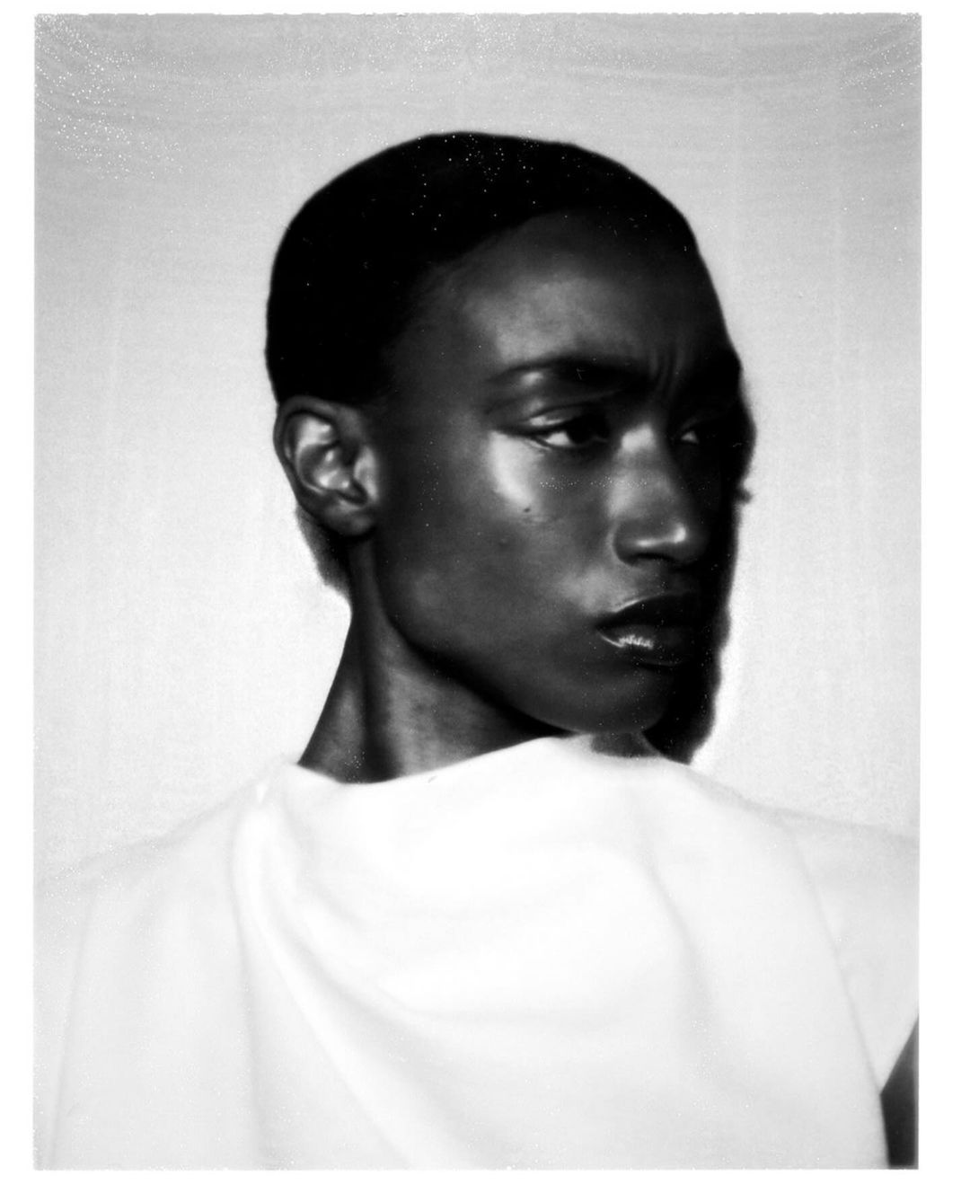 Black and white portrait of an African American female with dark hair and glossy lips, wearing a white top, against a plain backdrop.