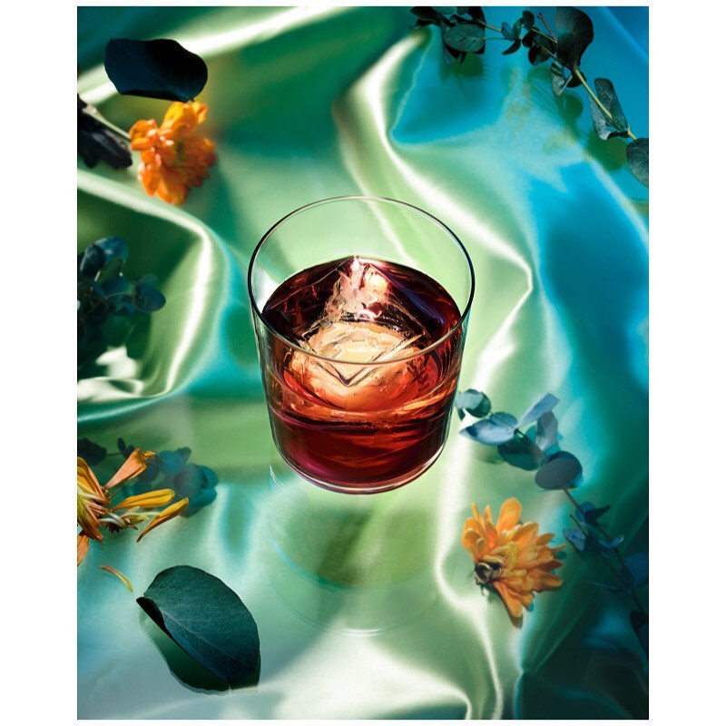 A stemmed glass of amber liquid with a large square ice cube sits on a reflective surface with scattered flowers and blue-green eucalyptus leaves.