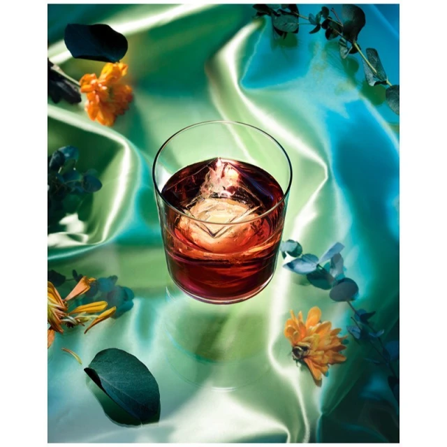 A stemmed glass of amber liquid with a large square ice cube sits on a reflective surface with scattered flowers and blue-green eucalyptus leaves.