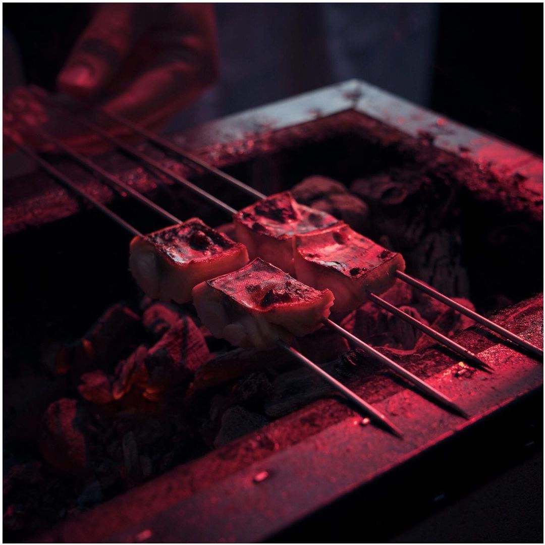 Three meat skewers cook over coals on a grill. The red light illuminates the grilling food and creates a warm atmosphere.
