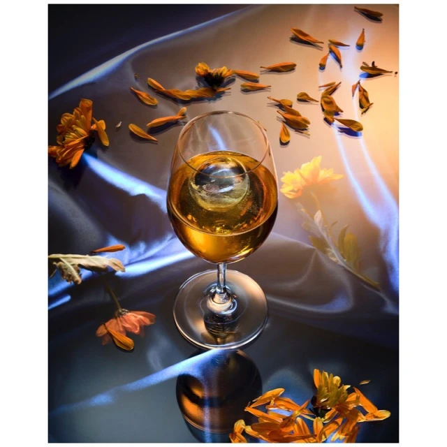 Wine glass filled with amber liquid and a sphere ice cube, adorned with scattered orange flower petals on a reflective surface.