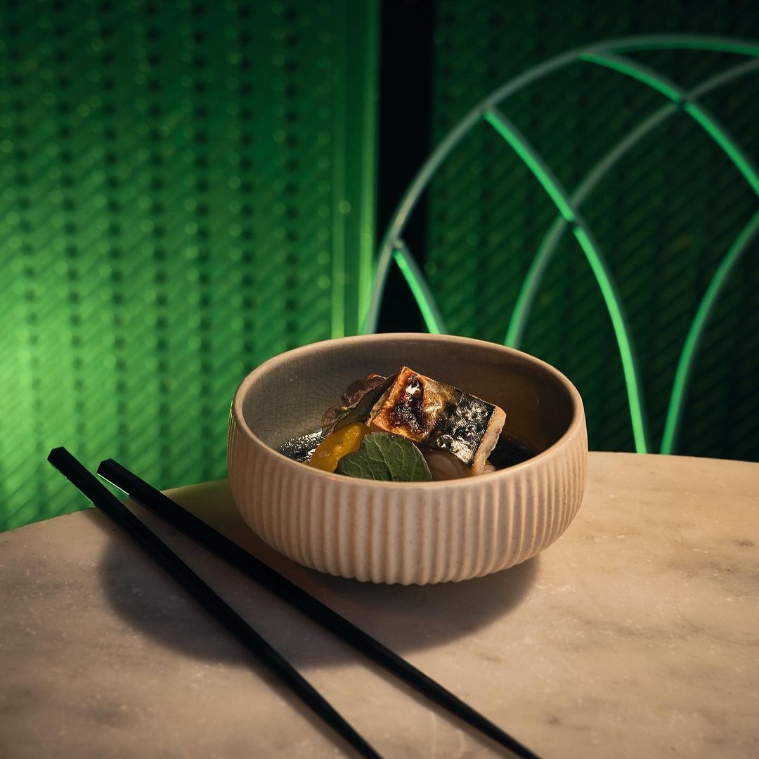 Close-up of a bowl of fish dish with chopsticks resting on a light marble table against a green patterned background.