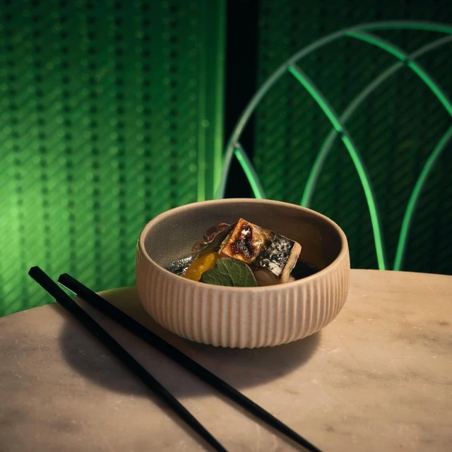 Close-up of a bowl of fish dish with chopsticks resting on a light marble table against a green patterned background.
