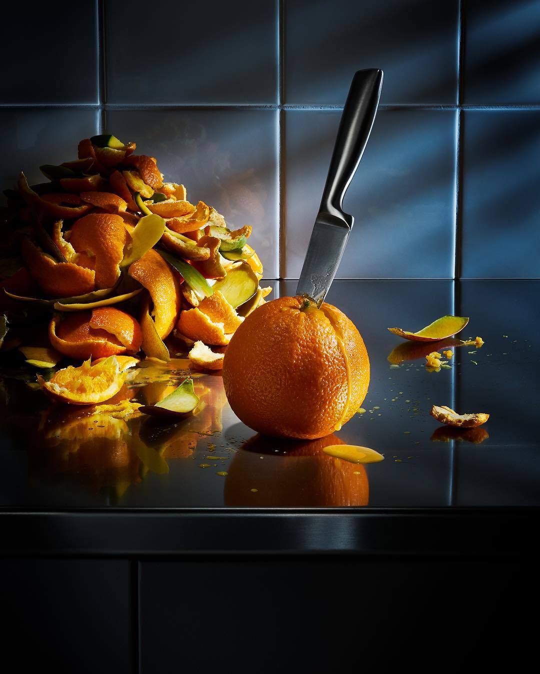 Orange and knife still life on reflective counter, with peels in background. Kitchen setting with tiled walls.