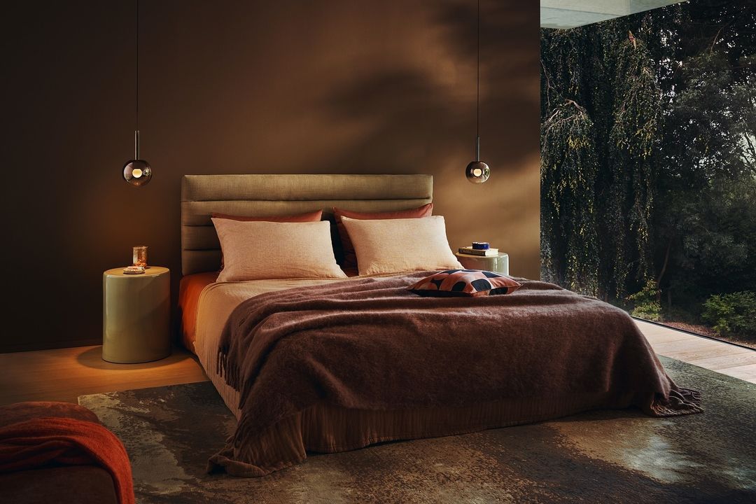 Modern bedroom interior with a made bed, brown walls, pendant lamps, and a large window showing green trees outside.