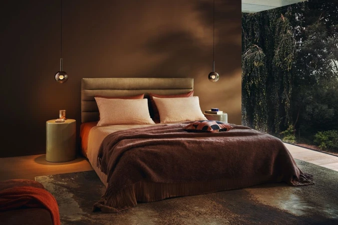 Modern bedroom interior with a made bed, brown walls, pendant lamps, and a large window showing green trees outside.