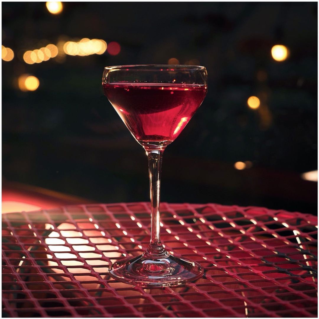Tall stemmed cocktail glass on a red wire surface, filled with a dark red drink with a blurred dark background and bokeh effect.