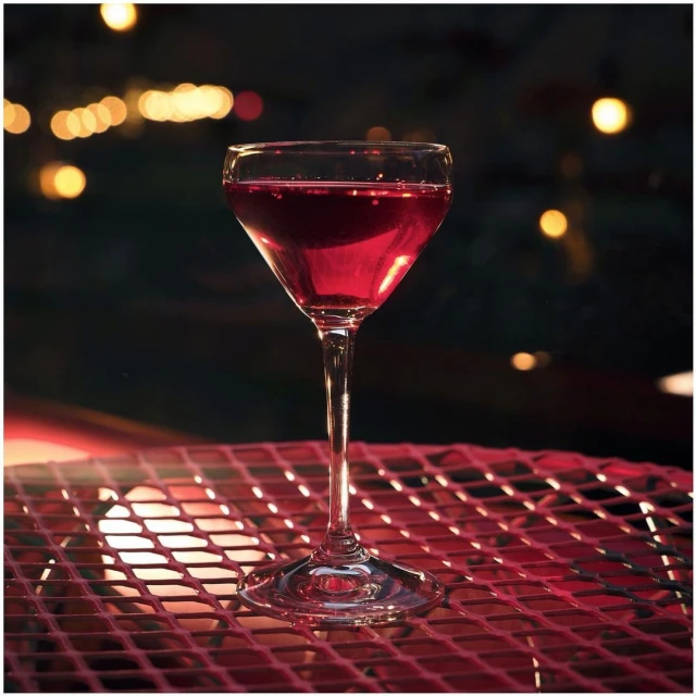 Tall stemmed cocktail glass on a red wire surface, filled with a dark red drink with a blurred dark background and bokeh effect.